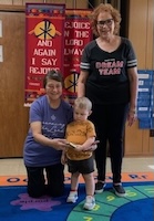 child holding a book with 2 adults looking on